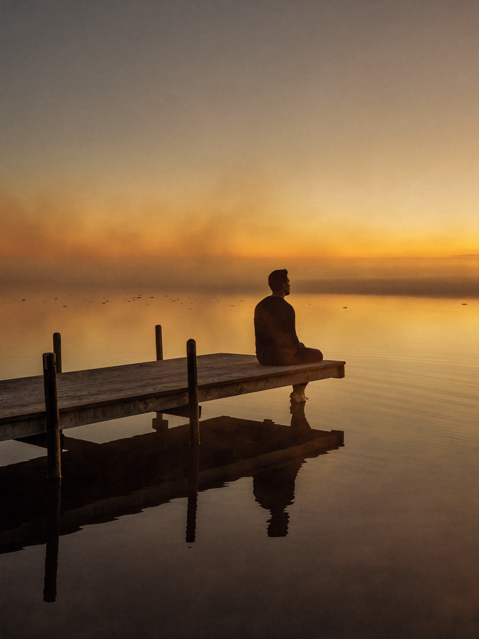 A man sitting in peaceful contemplation on a wooden dock at golden dawn, glassy water reflection, pure stillness