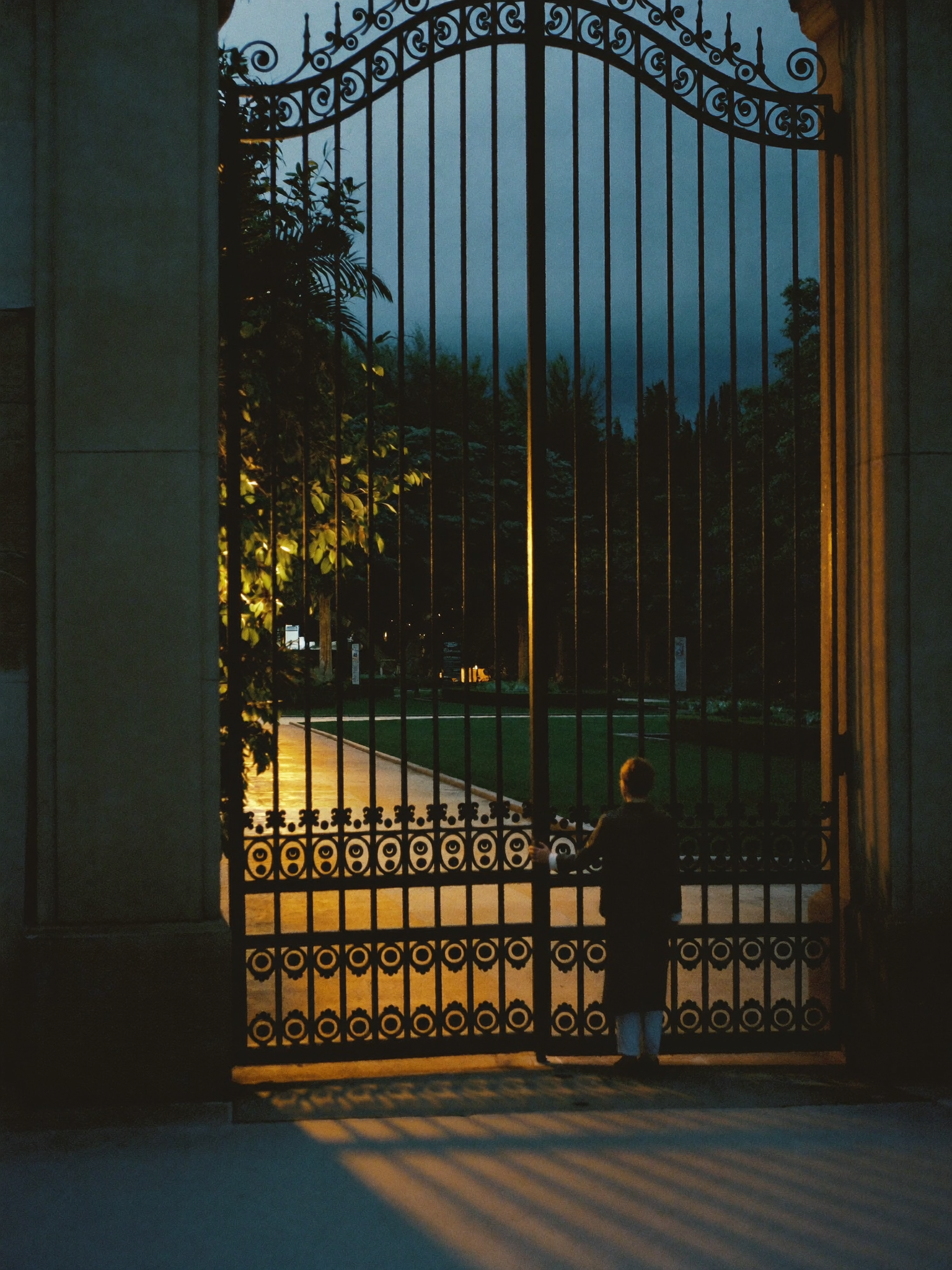 A lone figure standing before an ornate iron gate at dusk, the threshold between outside and a warm golden interior