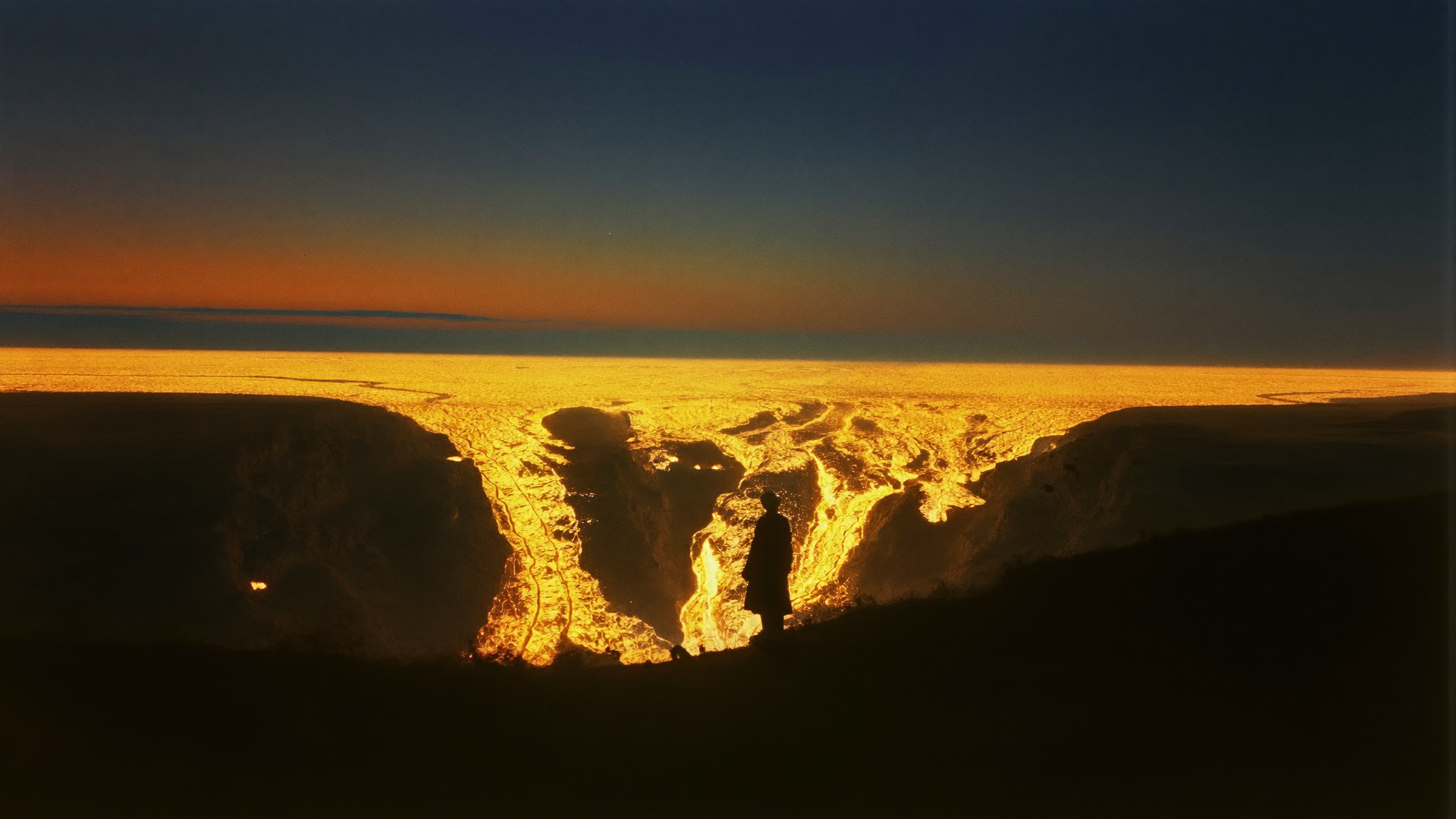 A silhouetted figure standing at the edge of a volcanic fissure at dusk, molten gold light splitting the earth at the horizon